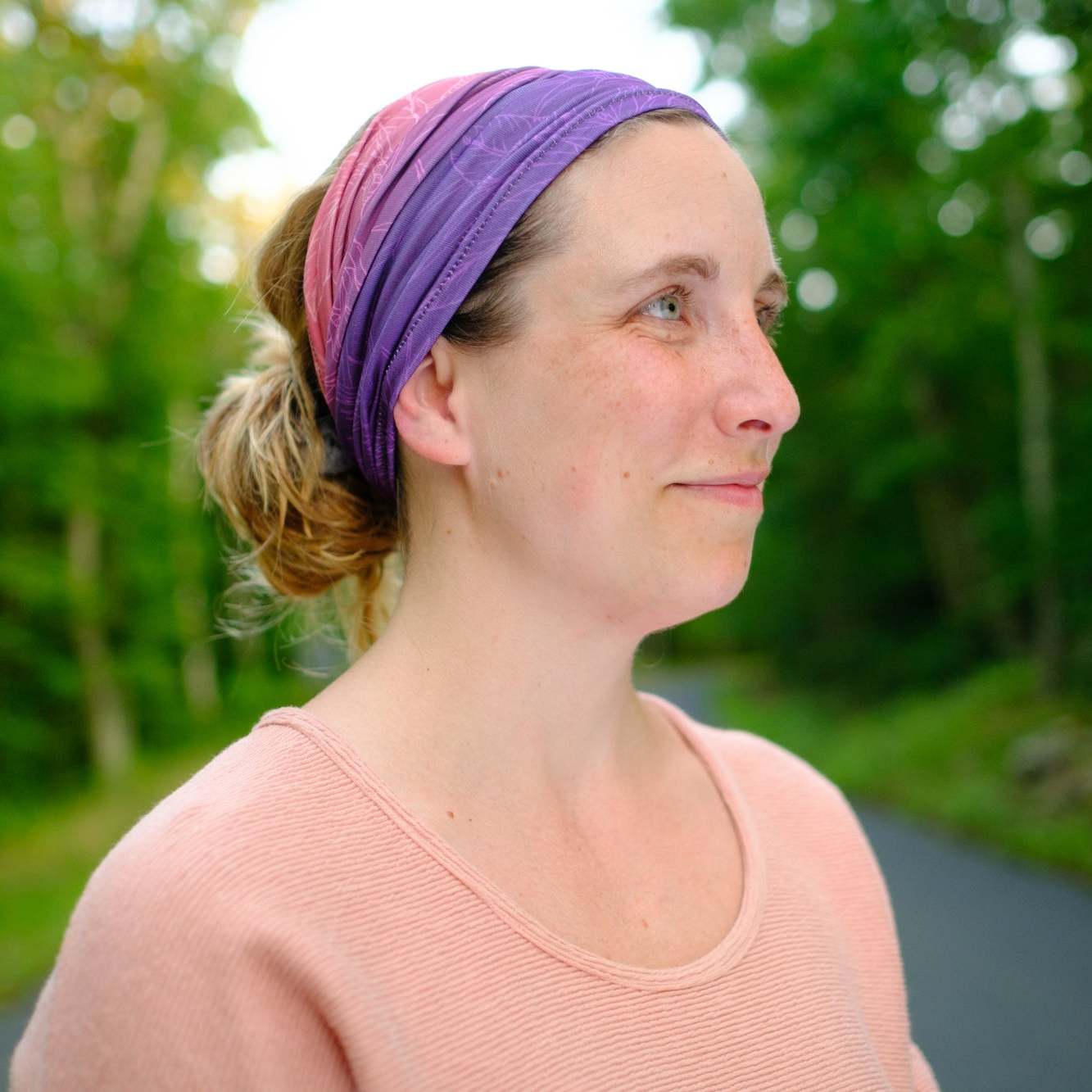 Woman wearing a purple neck gaiter as a headband with a blurred green outdoor background