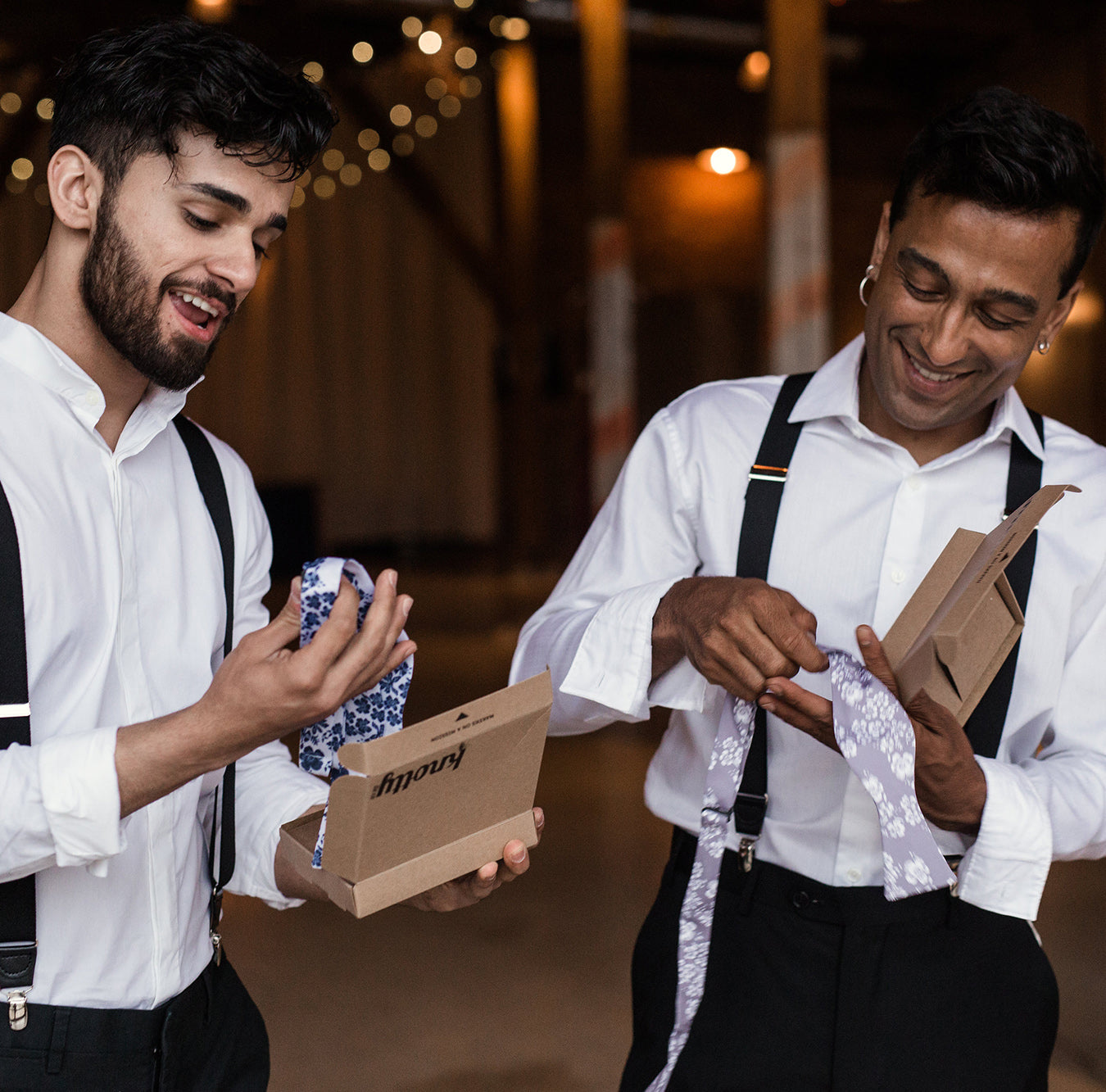 two men in white shirts and suspenders opening bow tie packages and smiling