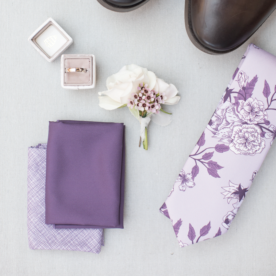 Purple pocket squares and floral necktie laying on table with flowers and ring box and shoes