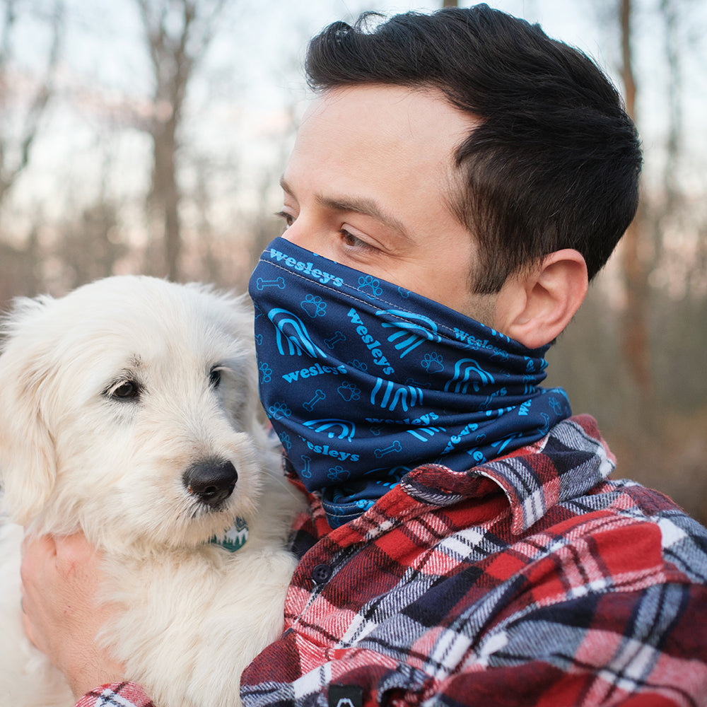 Man wearing a blue custom neck gaiter patterned face covering holding a white dog outdoors.