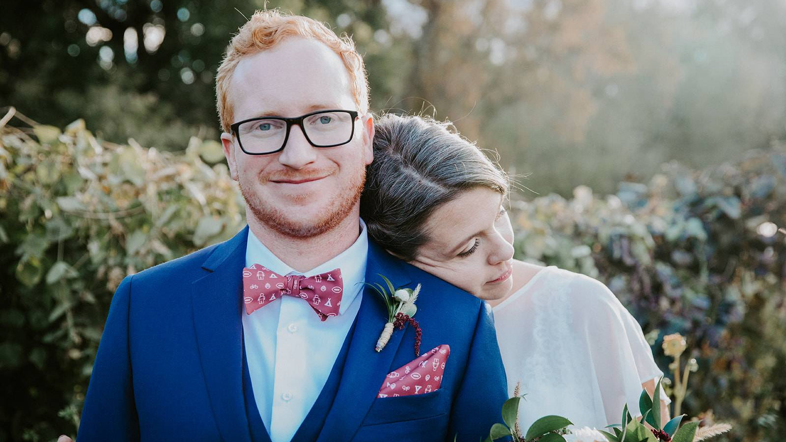 Man in a blue suit with a red bow tie and glasses, embracing a woman outdoors.