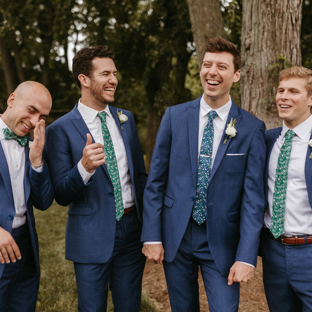 Four men in blue suits with green ties standing outdoors among trees.