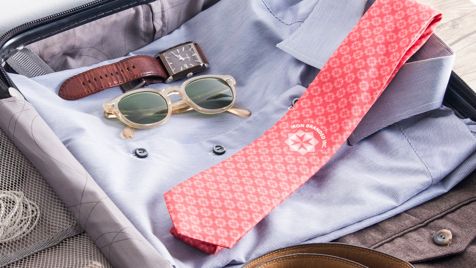 Open suitcase with shirt, sunglasses, watch, and red patterned tie.