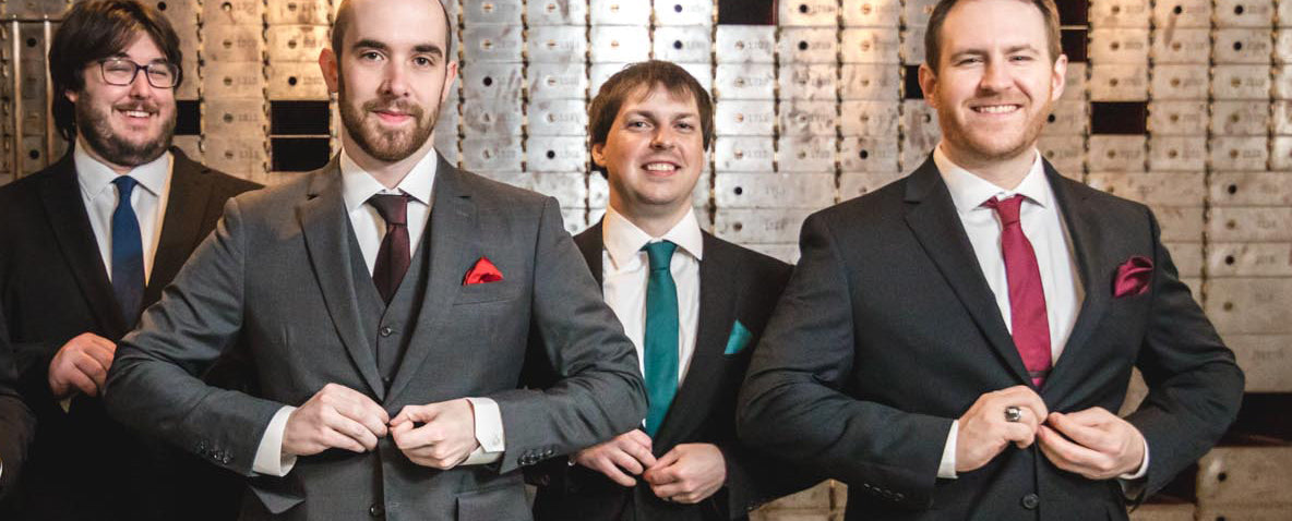 men in suits and ties and pocket squares standing in old bank
