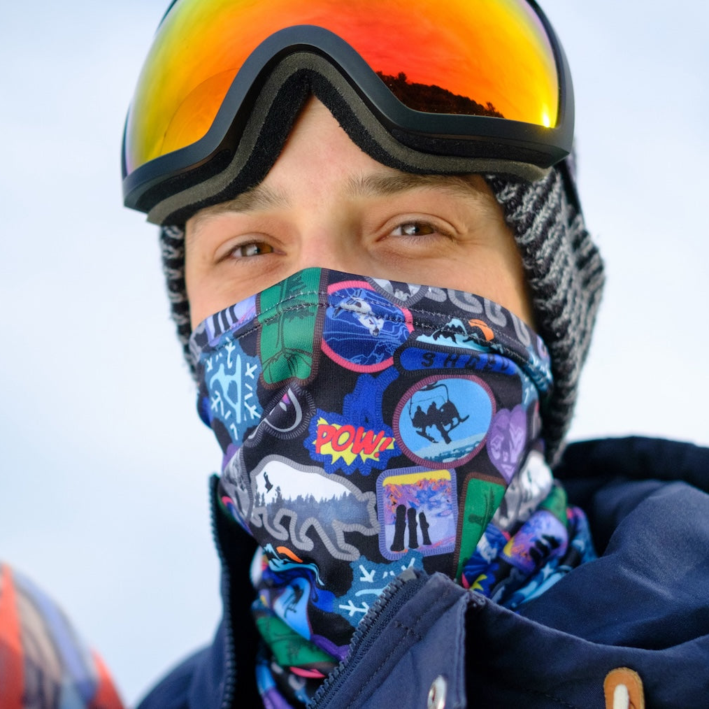 male snowboarder wearing a neck gaiter as a face covering and goggles