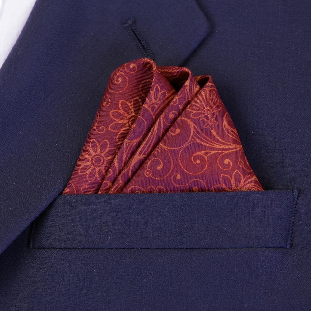 A wave folded patterned pocket square in magenta and orange tones, tucked into the breast pocket of a navy suit.
