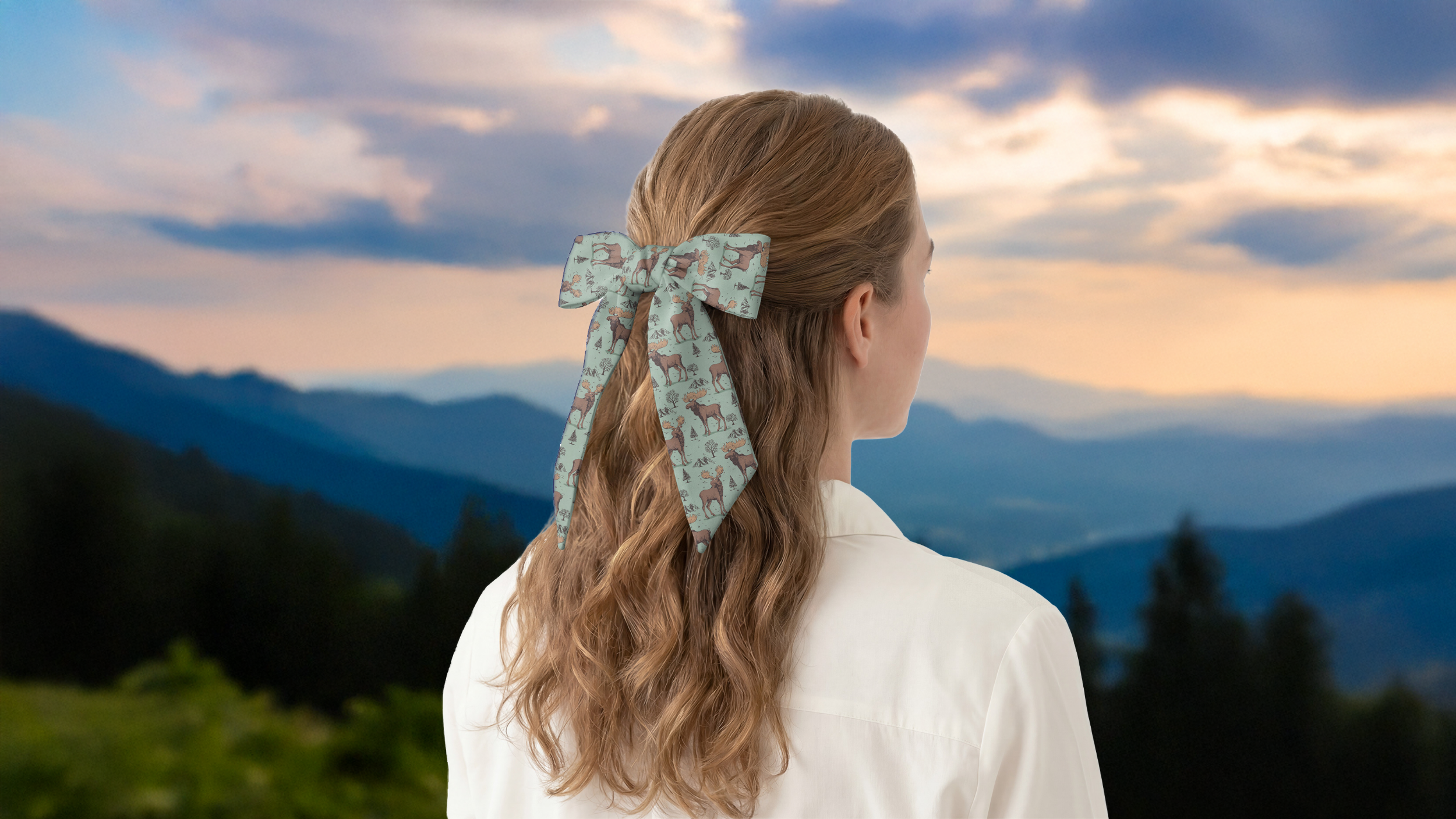 Person with a floral scarf in their hair standing against a mountainous landscape during sunset.