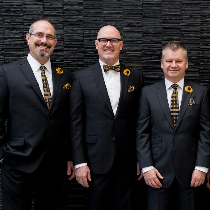Three men in suits with wedding bow ties and boutonnieres standing against a black textured wall.