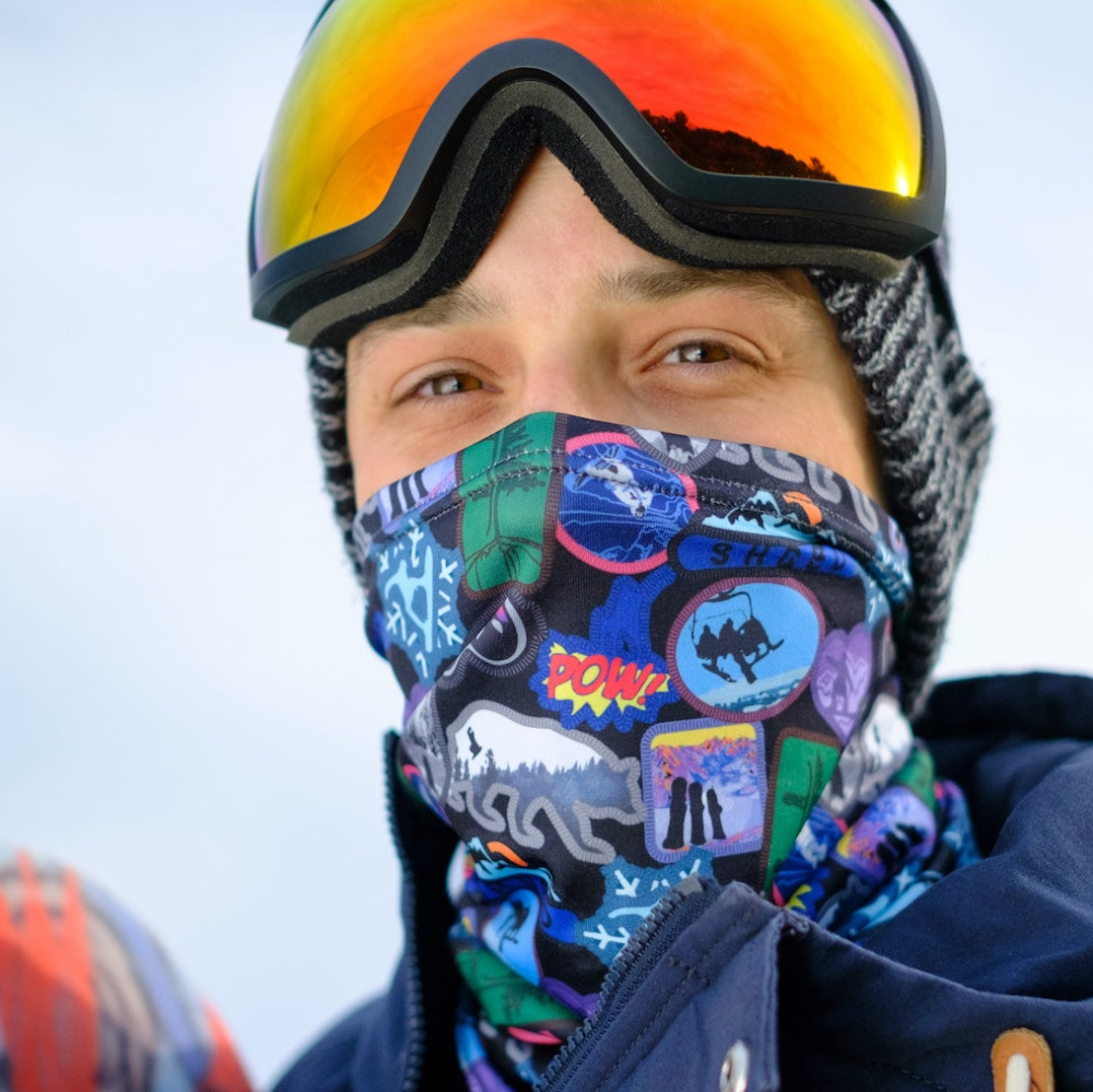 Person wearing a colorful patterned neck gaiter and reflective goggles against a light blue background