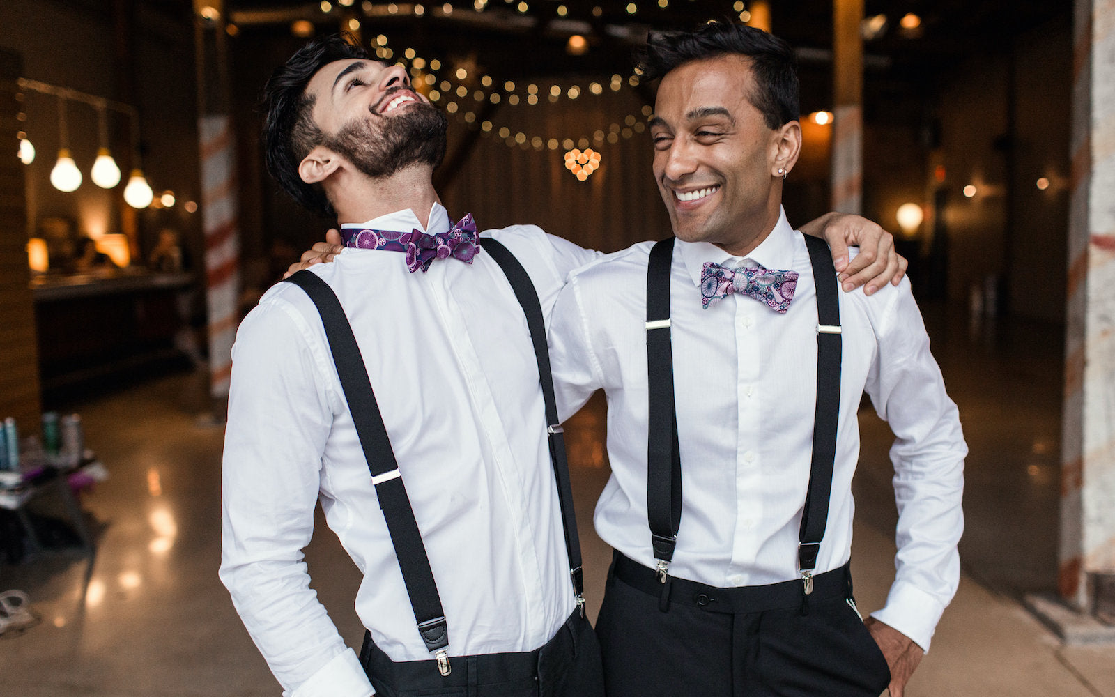 Two men in formal attire with suspenders and bow ties standing close together in a well-lit indoor setting.