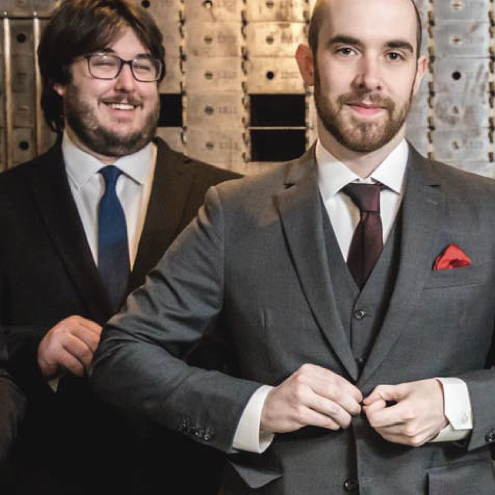 Two men in suits and pocket squares standing in front of a textured wall.