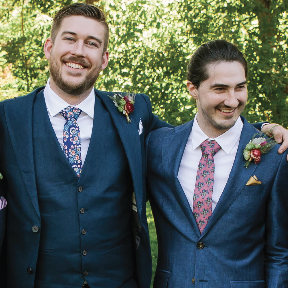Two men in suits with floral ties standing outdoors against a green background