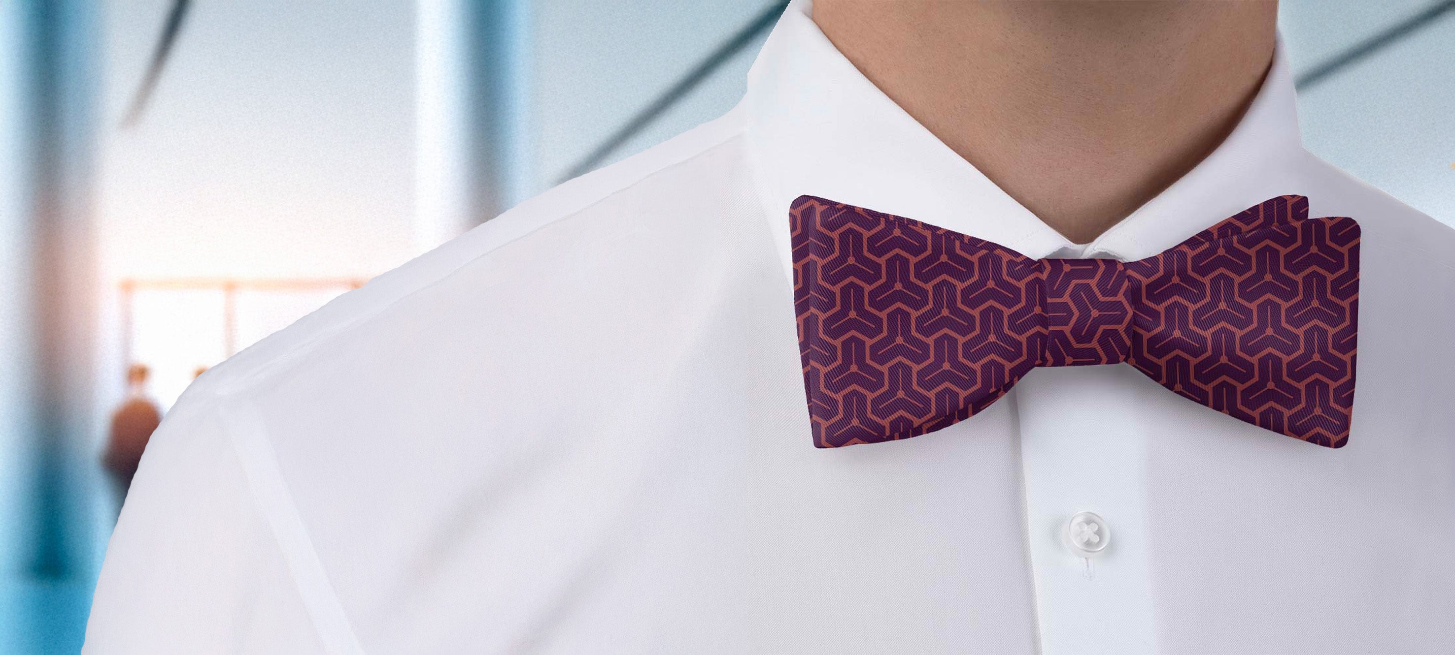 Man wearing a white shirt with a geometric patterned purple bow tie against a blurred indoor background