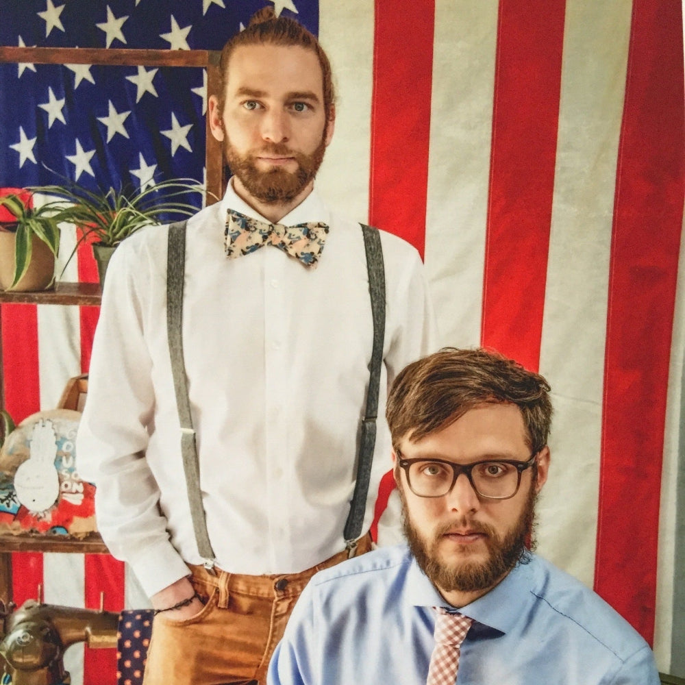 Two men posing in front of an American flag, one wearing suspenders and a bow tie, the other in a blue shirt with a tie