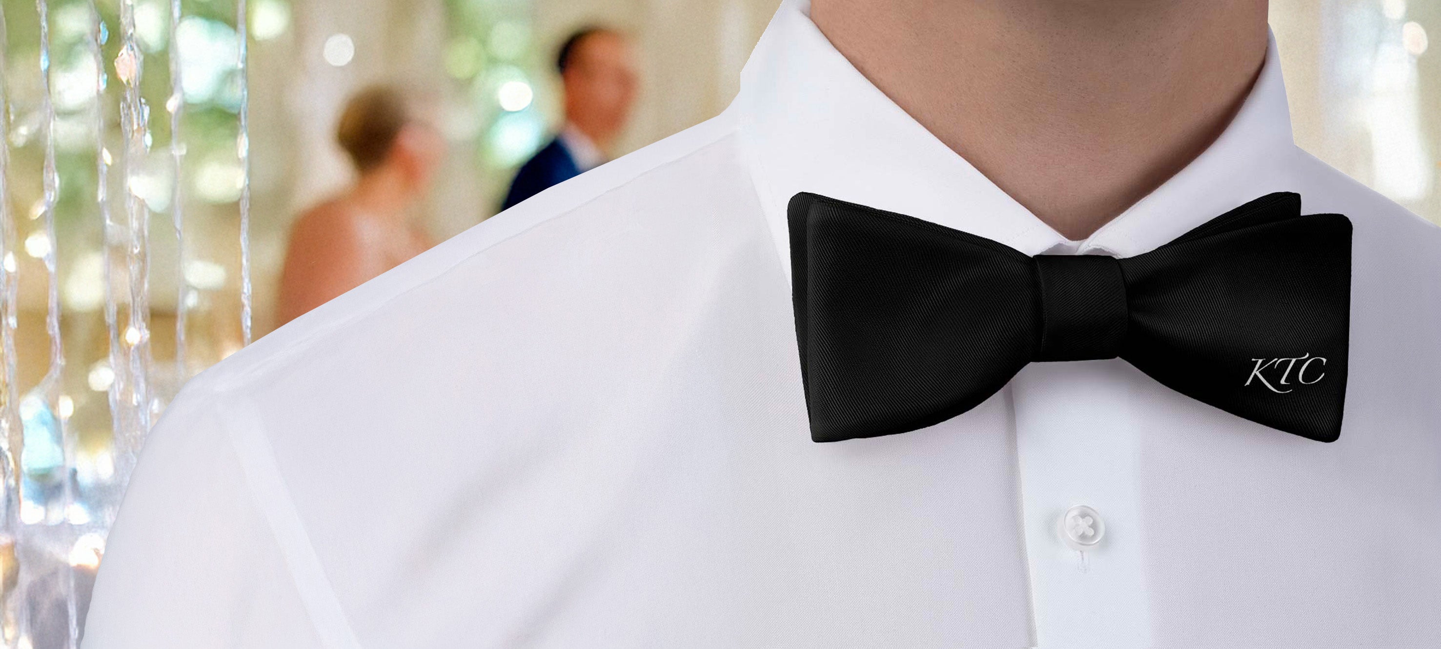 Person wearing a black monogram bow tie with 'KTC' initials in an elegant indoor wedding.