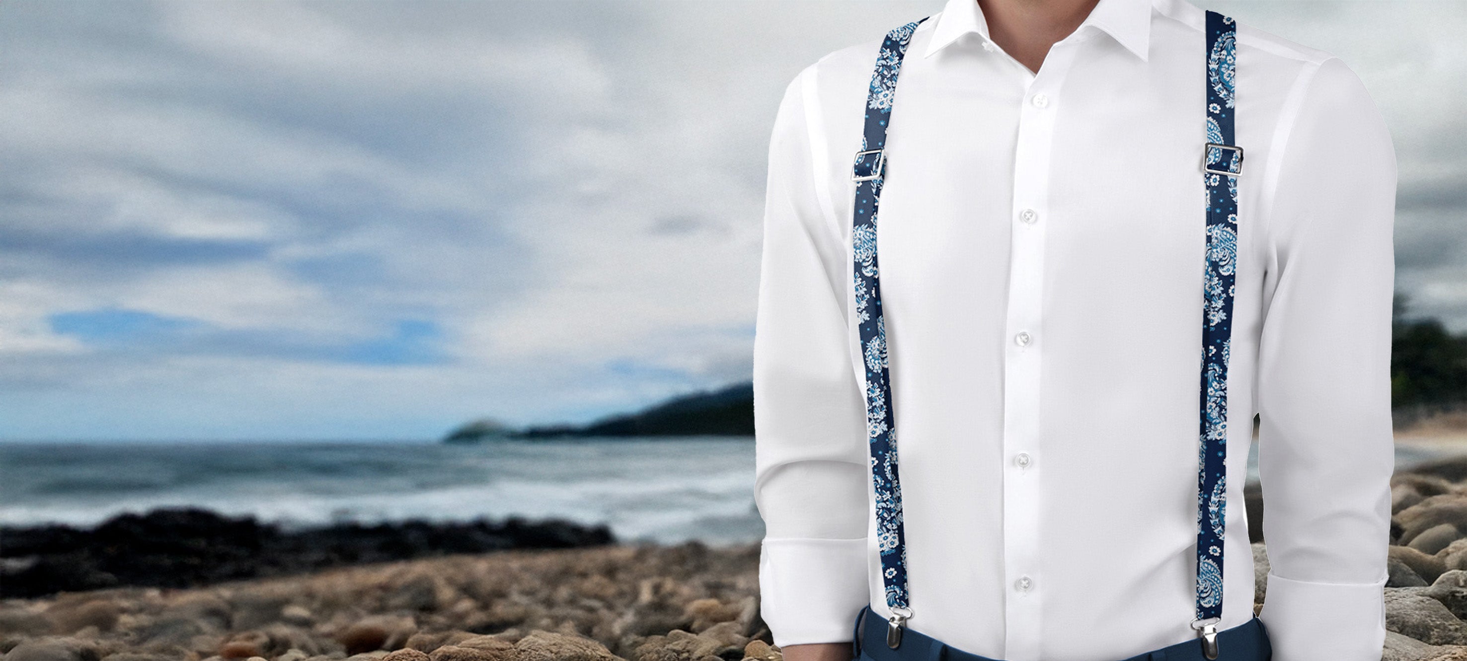 Person wearing a white shirt and blue paisley suspenders standing on a rocky beach with ocean and sky in the background