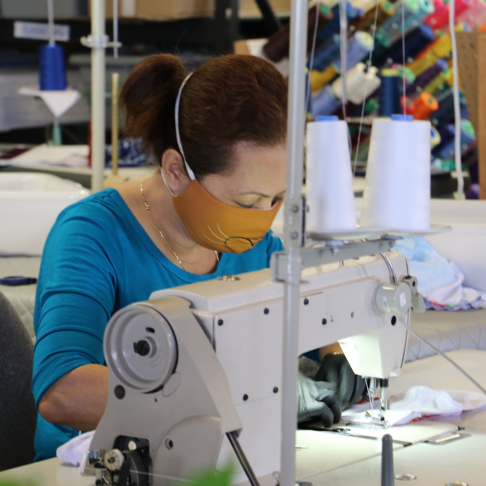 Person wearing a face mask working on a sewing machine in a factory setting.