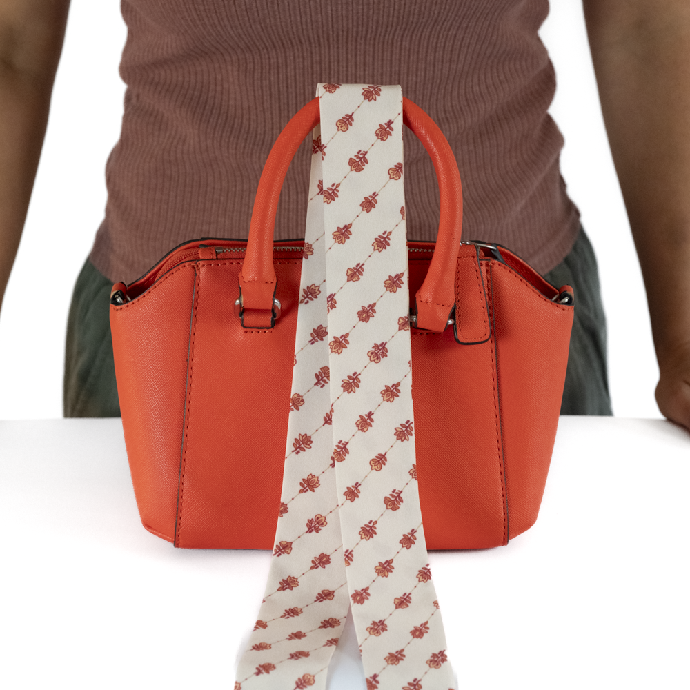 An orange leather handbag with a long cream-colored skinny scarf draped over it.