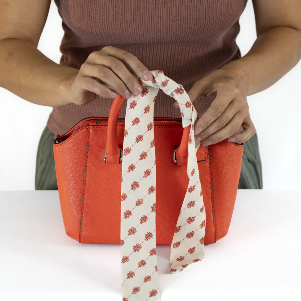 A person wrapping a cream-colored fabric scarf around the handle of a bright orange handbag.