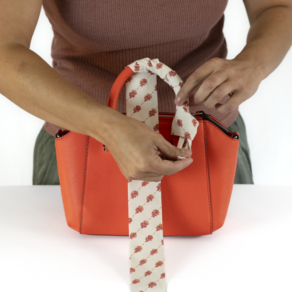 A person's hands are shown tying a knot at the end of a cream-colored scarf wrapped around the handle of a bright orange handbag.