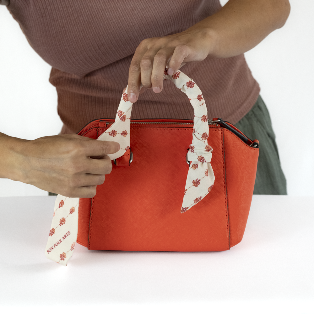 A person wrapping a cream-colored fabric scarf around the other side of the handle of a bright orange handbag.