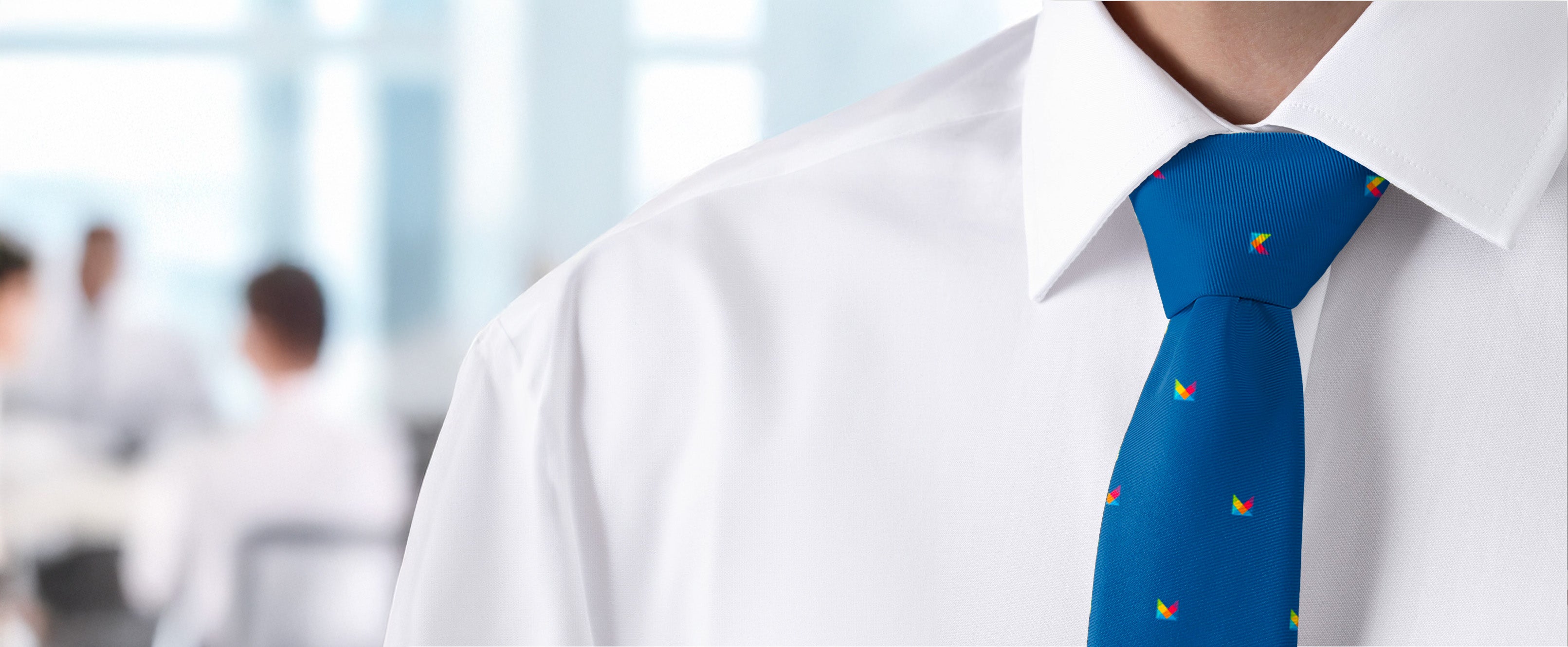 Close-up of a person wearing a white shirt and blue tie with colorful logo patterns, blurred office background.