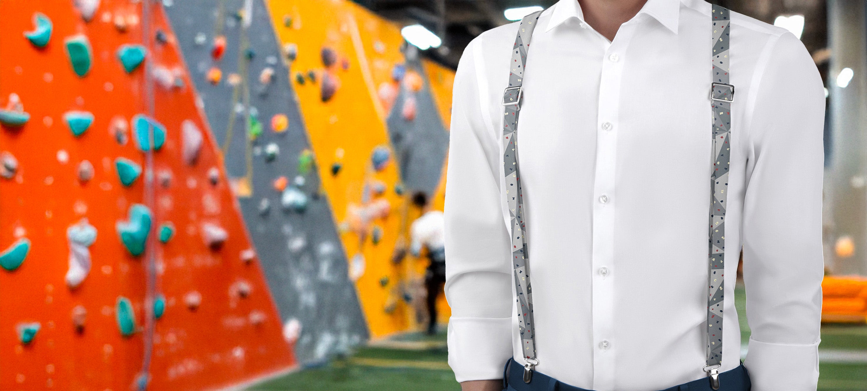 Person wearing a white shirt and climbing suspenders standing in front of a colorful climbing wall.