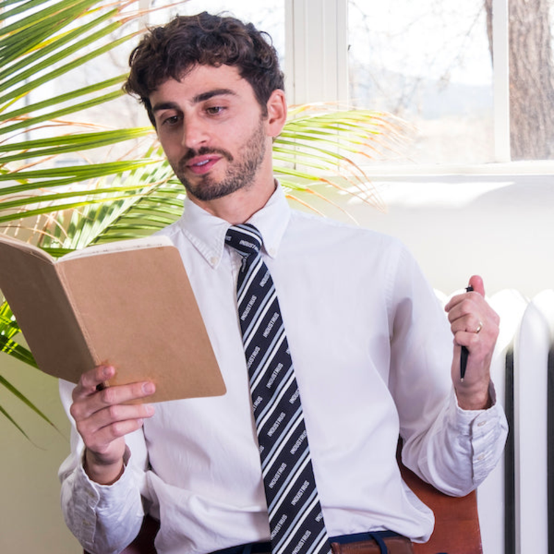 Man in a white shirt and tie holding a notebook and pen, sitting in a bright room with plants.