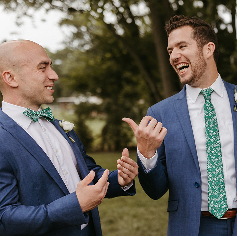 Two men in suits with green bow ties and floral boutonnieres are laughing outdoors.