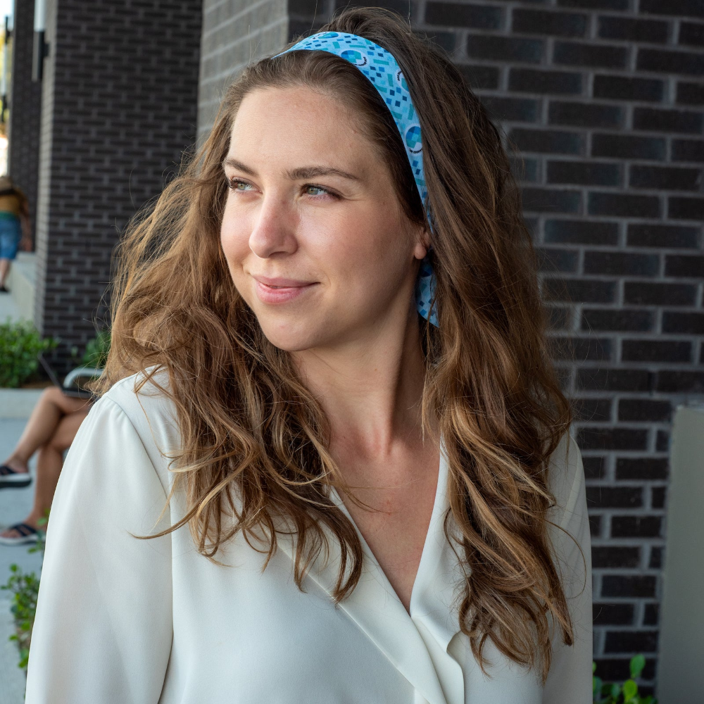 Woman with a blue hair scarf and white shirt standing against a brick wall.