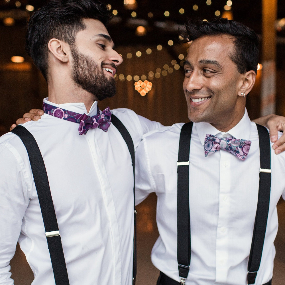 Two men wearing white shirts, black suspenders, and colorful bow ties.